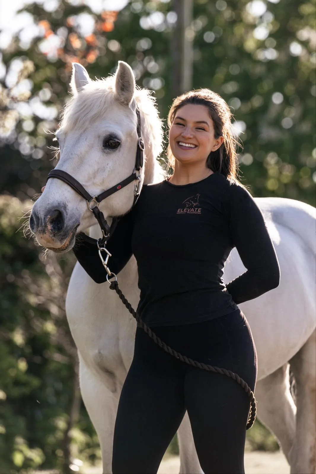 Elena smiling alongside a white horse, wearing Elevate Vet Physio branded top